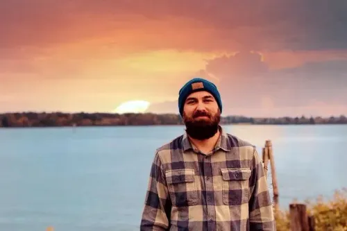 رجل يقف أمام بحيرة تحت سماء غائمة، يرتدي قبعة زرقاء وقميص مخطط. تعكس الصورة التحديات التي يواجهها العمال الفيدراليون بعد فقدان وظائفهم.