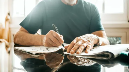 رجل مسن يحل لغز الكلمات المتقاطعة على طاولة، مما يعكس أهمية الأنشطة العقلية النشطة في الحفاظ على صحة الدماغ.