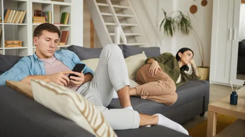 شخصان يجلسان على أريكة في غرفة معيشة، الرجل مشغول بهاتفه بينما المرأة تبدو حزينة ومفكرة، مما يعكس حالة الطلاق الصامت.
