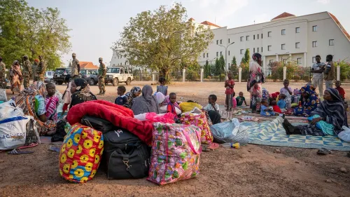 عائلات نازحة في جوبا بجنوب السودان، تجلس مع حقائب وأمتعة، بينما توجد قوات أمنية في الخلفية.