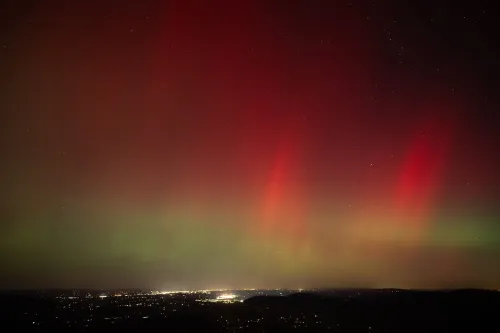شفق قطبي ملون يتراقص في السماء ليلاً فوق منطقة حضرية، مع أضواء حمراء وخضراء، نتيجة لعاصفة إشعاعية شمسية قوية.