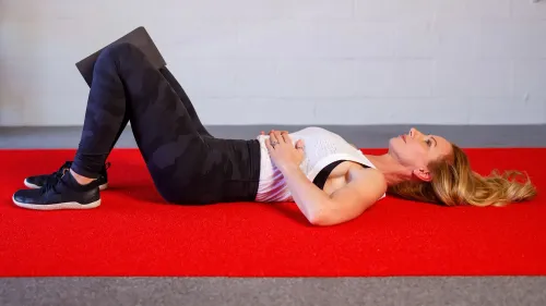 امرأة مستلقية على سجادة حمراء، تمسك بيدها على ضلوعها السفلية، وتقوم بتقنية تنفس موجهة لتخفيف التوتر واستعادة المحاذاة.