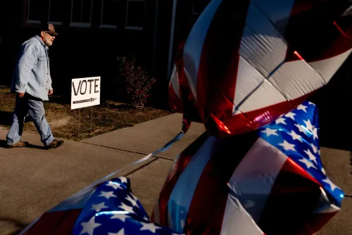 رجل يسير نحو لافتة "صوت" بينما تتدلى بالونات مزينة بالعلم الأمريكي، مما يعكس أجواء الانتخابات.
