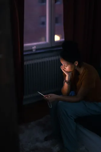 امرأة شابة تجلس في غرفة ذات إضاءة خافتة، تنظر بقلق إلى هاتفها المحمول، مما يعكس تأثير وسائل التواصل الاجتماعي على رفاهيتها.
