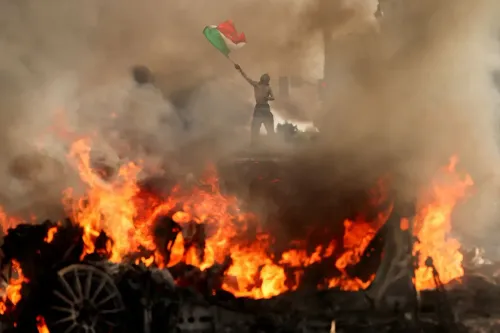 متظاهر يحمل علم فلسطين وسط دخان ونيران في احتجاجات، مما يعكس التوترات الاجتماعية والسياسية الحالية.