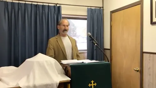 قس يتحدث في تجمع محلي، يرتدي بدلة، مع وجود ميكروفون أمامه وغطاء طاولة في الخلفية. تتعلق الصورة بانتخابات مجلس الشيوخ في نبراسكا.