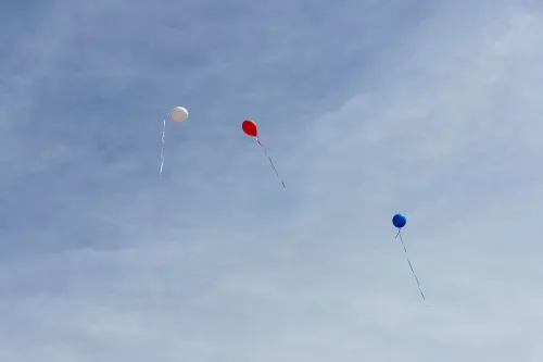 ثلاثة بالونات ملونة (أحمر، أزرق، أبيض) تطفو في السماء الزرقاء، تعكس شعور الأمل والتحديات التي يواجهها المهاجرون.