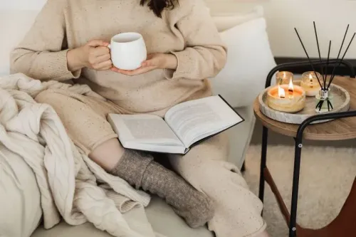 امرأة ترتدي ملابس مريحة تحمل كوبًا من المشروبات الساخنة، تجلس بجانب كتاب مفتوح على طاولة صغيرة، مع شموع ومكان مريح للقراءة.