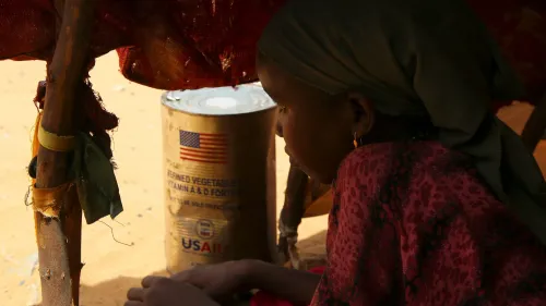 طفلة تجلس بجوار علبة غذائية تحمل شعار الوكالة الأمريكية للتنمية الدولية (USAID)، تعكس تأثير إنهاء المساعدات الإنسانية.