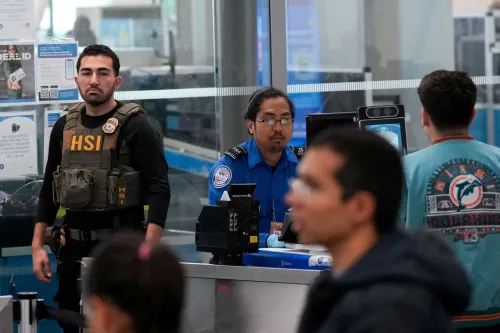 موظفان من إدارة أمن النقل يقفان في نقطة تفتيش بمطار، مع وجود مسافرين في الخلفية، وسط توتر حول تمويل الوزارة.