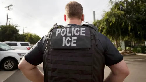 ضابط إنفاذ قانون يرتدي سترة مكتوب عليها \"شرطة ICE\" في موقع خارجي، مما يعكس تغييرات السياسة في اعتقال المهاجرين.