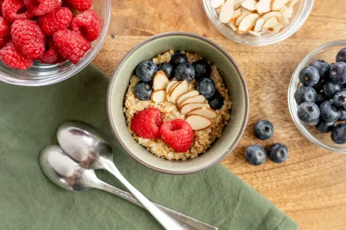 وعاء من الشوفان مع توت العليق والتوت الأزرق وشرائح اللوز، محاطًا بأوعية من الفواكه الطازجة، على سطح خشبي.