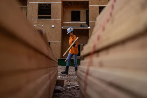 شابة تحمل قطعة خشب في موقع بناء، مع وجود صفوف من الخشب في المقدمة وخلفية تظهر المبنى قيد الإنشاء.