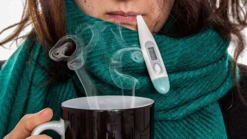امرأة ترتدي وشاحًا أخضر، تحمل كوبًا بخاريًا، ومقياس حرارة في فمها، تعكس أعراض الإنفلونزا في موسمها الحالي.