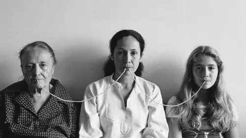 صورة بالأبيض والأسود لثلاث نساء من أجيال مختلفة يجلسن معًا، ويمتد خيط بين أفواههن، مما يعكس موضوع التواصل بين الأجيال.