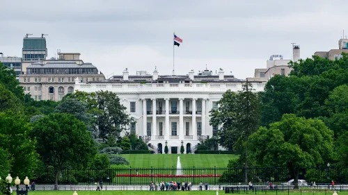 البيت الأبيض يظهر في صورة تحت سماء غائمة، مع العلم الأمريكي يرفرف فوقه، بينما يتجمع عدد من الأشخاص في الحديقة.