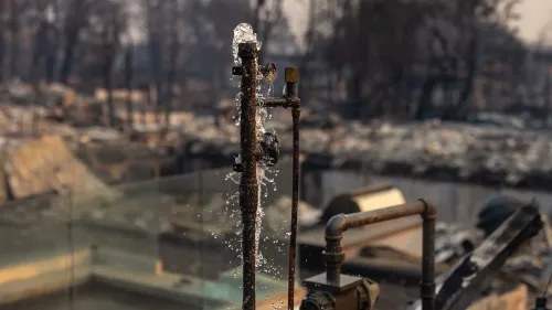 تظهر الصورة أنبوب مياه متضرر يتدفق منه الماء، مع خلفية مدمرة بسبب حرائق الغابات في لوس أنجلوس، مما يبرز خطر تلوث مياه الشرب.