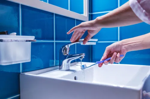 يد تظهر يدًا تمسك بفرشاة أسنان بالقرب من حوض غسيل، بينما تتدفق المياه من الصنبور الأزرق، مما يعكس روتين العناية بالفم.
