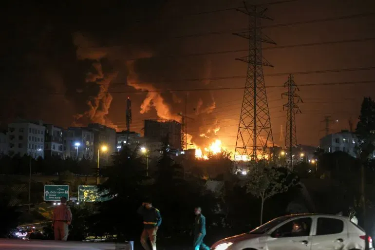 دخان كثيف يتصاعد من مناطق سكنية في طهران بعد هجمات إسرائيلية، مع وجود أشخاص يتجولون في الشوارع ليلاً.