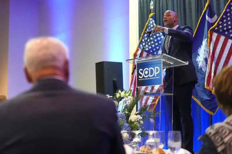 خطيب يتحدث في حفل عشاء لحزب الديمقراطيين في ساوث كارولينا، مع لافتة الحزب وخلفية من الأعلام الأمريكية، وسط تفاعل الحضور.