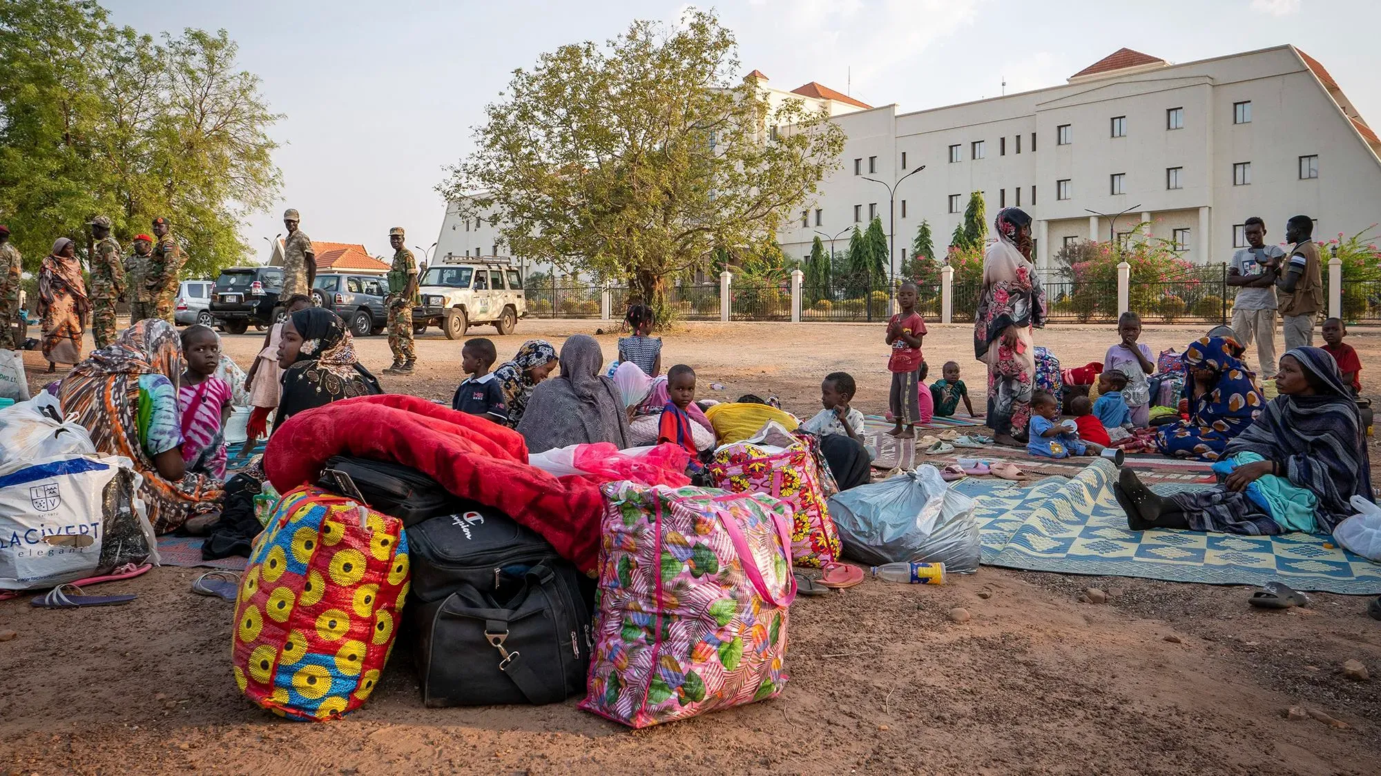 عائلات نازحة في جوبا بجنوب السودان، تجلس مع حقائب وأمتعة، بينما توجد قوات أمنية في الخلفية.