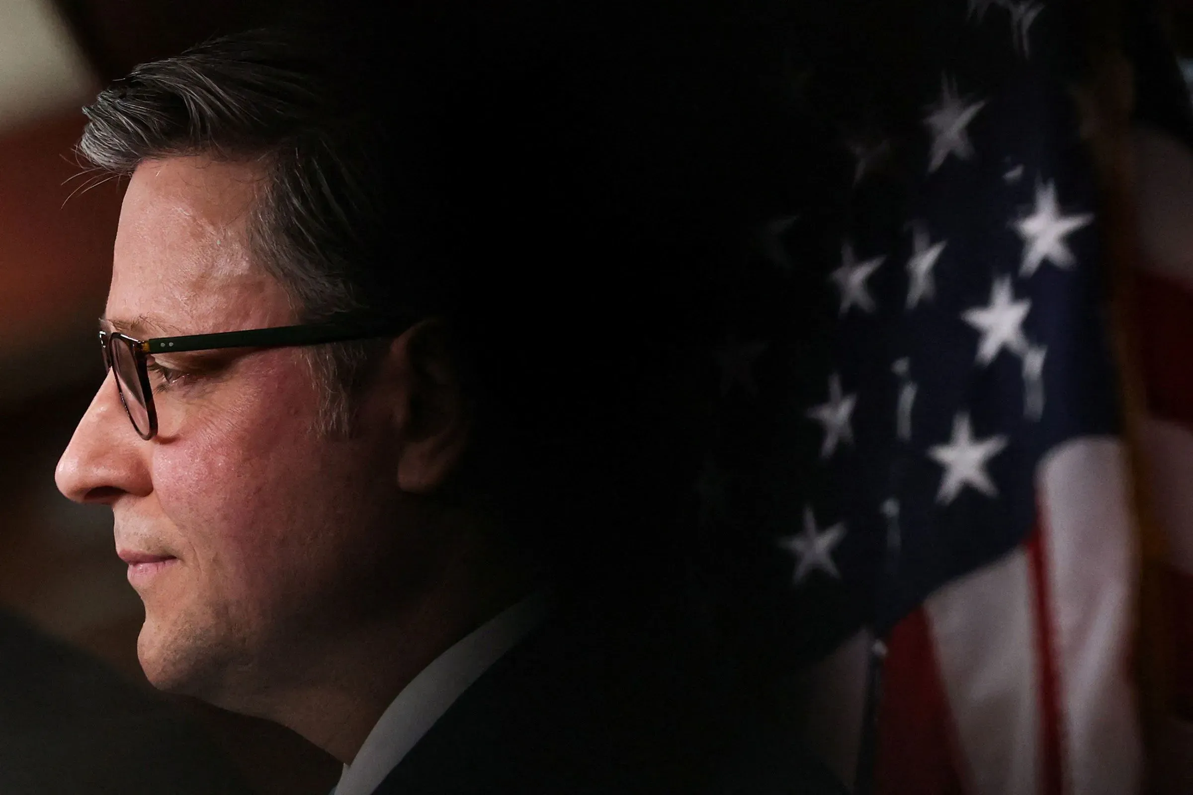 مايك جونسون، رئيس مجلس النواب الأمريكي، يظهر في صورة جانبية مع العلم الأمريكي خلفه، مع التركيز على تعبير وجهه الجاد.