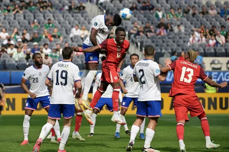 تظهر الصورة لحظة تنافسية في مباراة كرة قدم بين الولايات المتحدة وكندا، حيث يقف لاعبون من الفريقين في مواجهة بعضهم، مع محاولة أحد اللاعبين القفز لتهيئة الكرة.