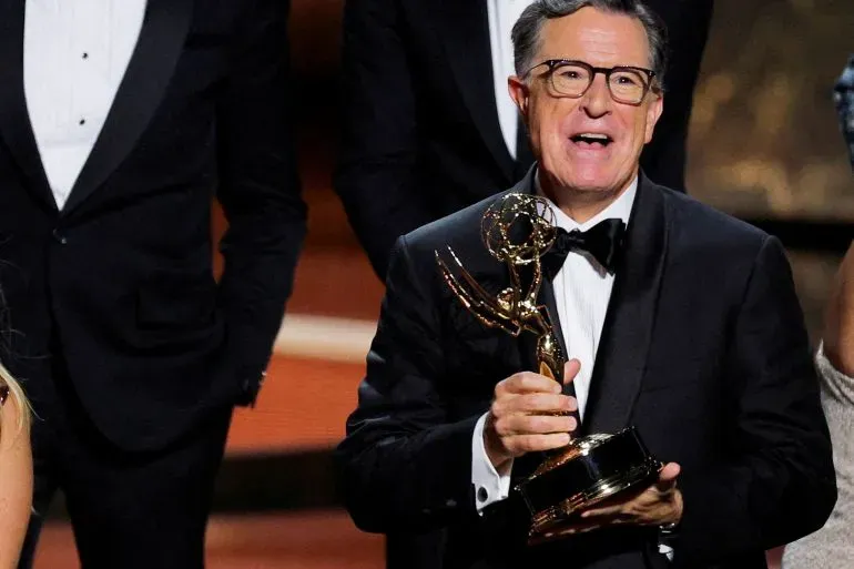 ستيفن كولبير يحمل جائزة إيمي، مبتسمًا، بينما يرتدي بدلة رسمية. خلفه يظهر عدد من الأشخاص في ملابس أنيقة، مما يعكس أجواء احتفالية.