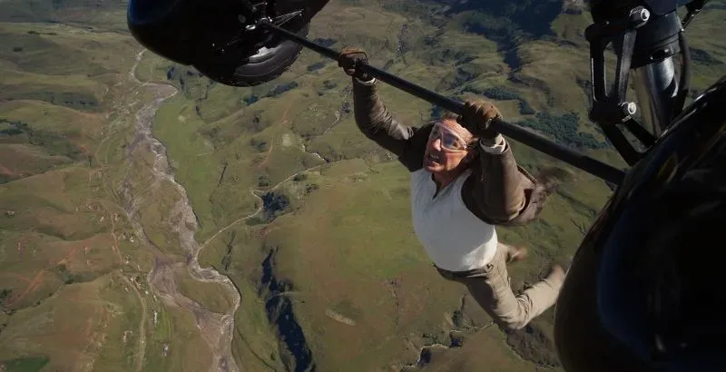 مشهد مثير من فيلم "Mission: Impossible" يظهر شخصية تتدلى من طائرة هليكوبتر فوق منظر طبيعي جبلي، مما يعكس أجواء الإثارة والتشويق.