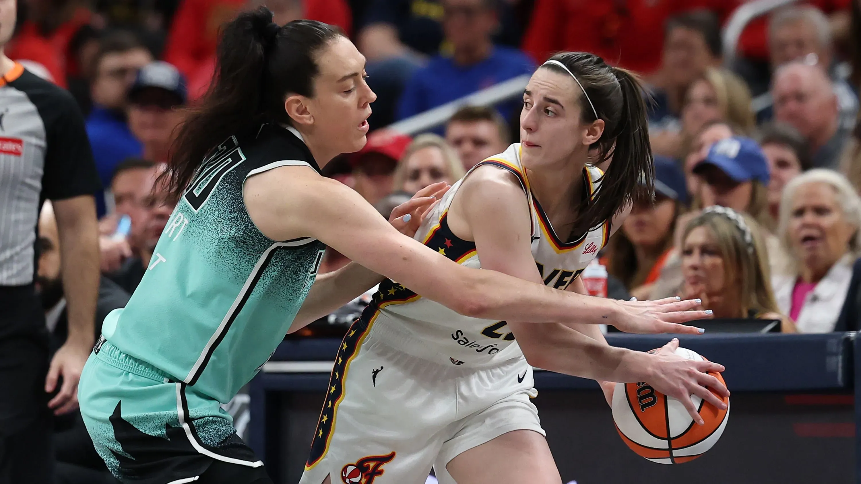 مباراة كرة سلة نسائية بين لاعبتين، حيث تحاول إحداهما استعادة الكرة بينما الأخرى تدافع عنها، مع جمهور في الخلفية.