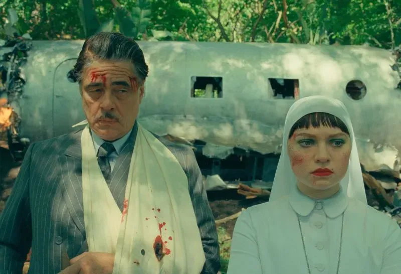 مشهد من فيلم "المخطط الفينيقي" يظهر شخصيتين رئيسيتين في غابة، مع خلفية حطام طائرة، تعكس أجواء الإثارة والغموض.