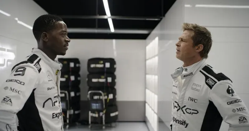مشهد من فيلم "F1" يظهر سائقين يتبادلان النظرات في حلبة السباق، مع خلفية من الإطارات والمعدات، مما يعكس أجواء الإثارة والتنافس في سباقات الفورمولا 1.
