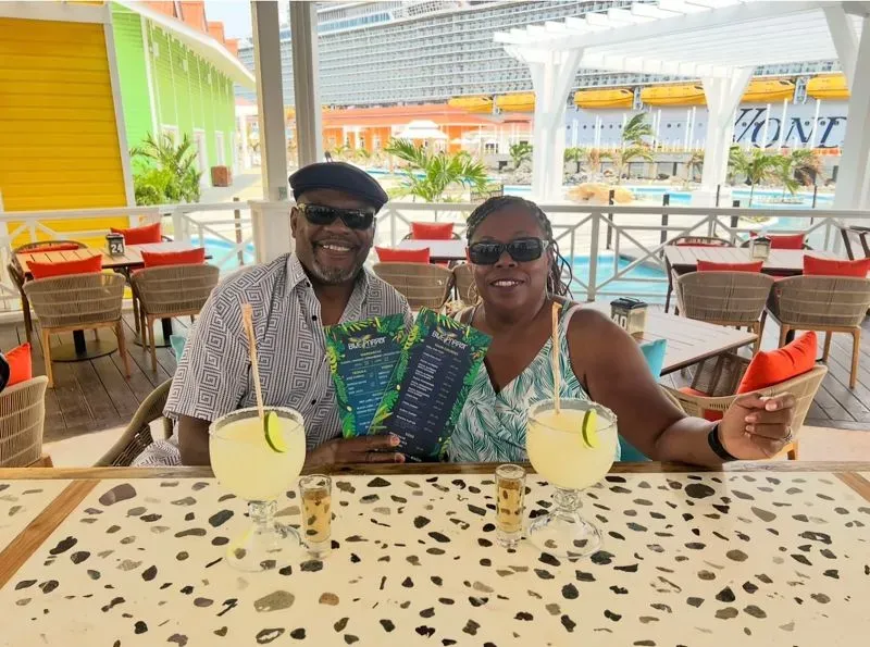 زوجان يستمتعان بوقتهما في بار على متن سفينة سياحية، يحملان قائمة الطعام أمامهم مع خلفية ملونة تضم مسابح وأسطح زلاجات مائية.