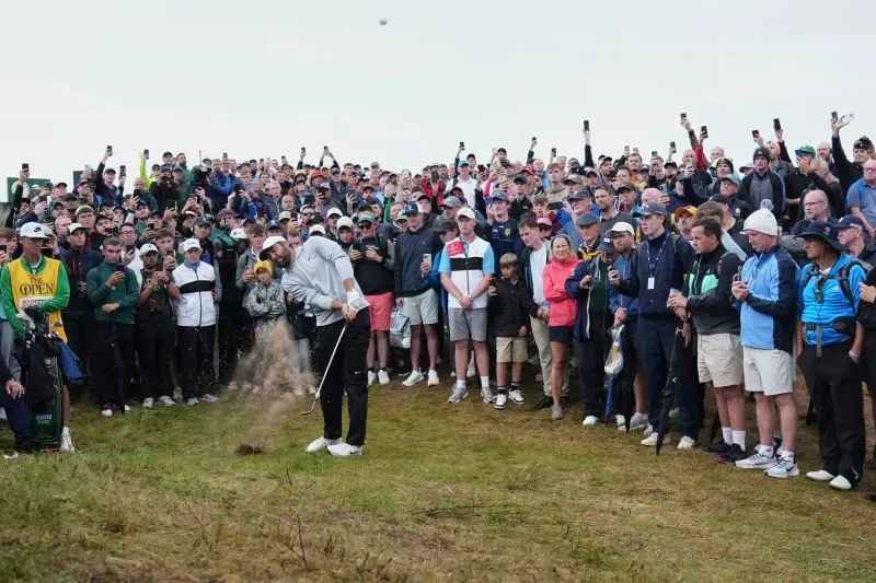لاعب جولف يضرب الكرة من الرمال أمام حشد كبير من المشجعين، مع هواتفهم المحمولة في الهواء، في أجواء مشمسة وغائمة.