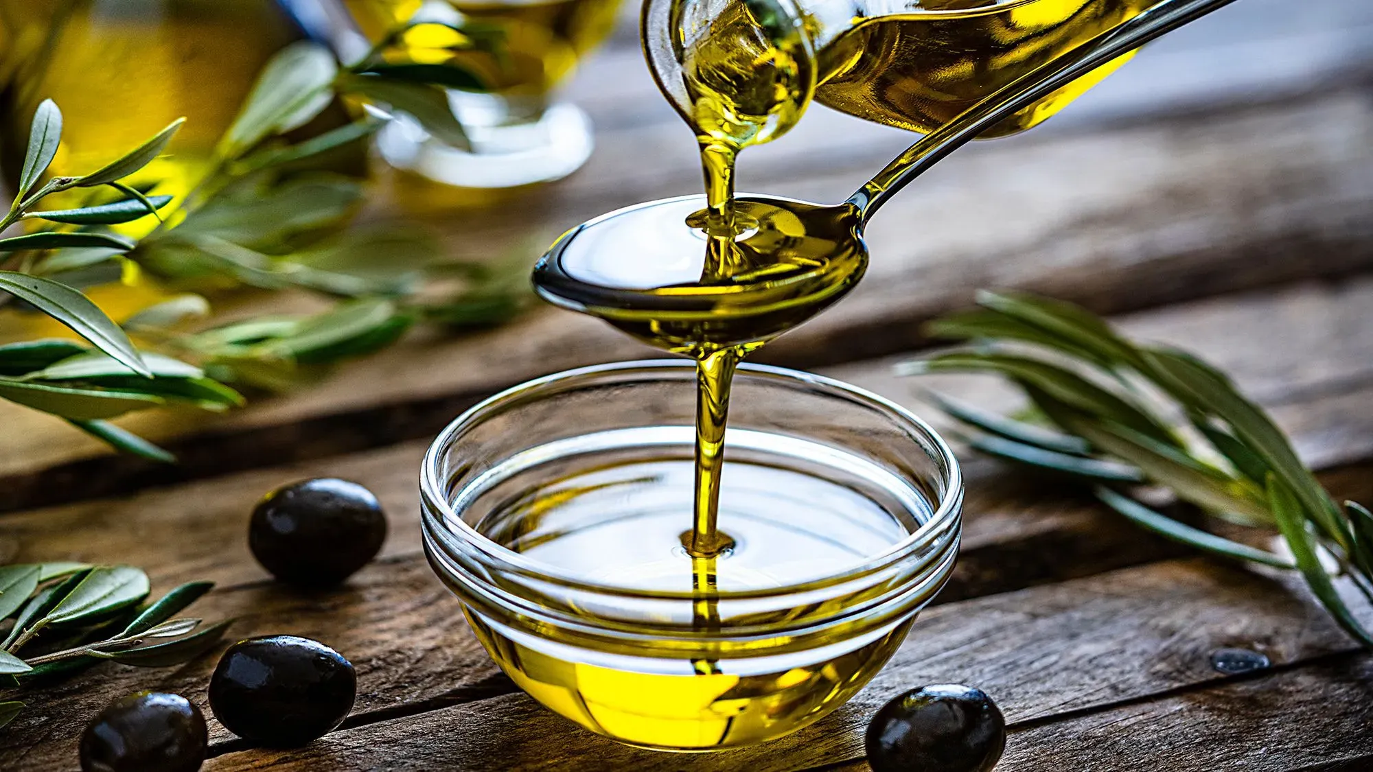 زيت زيتون يُسكب في وعاء زجاجي، مع زيتون وأوراق زيتون على سطح خشبي، مما يُبرز فوائد الزيوت النباتية الصحية.