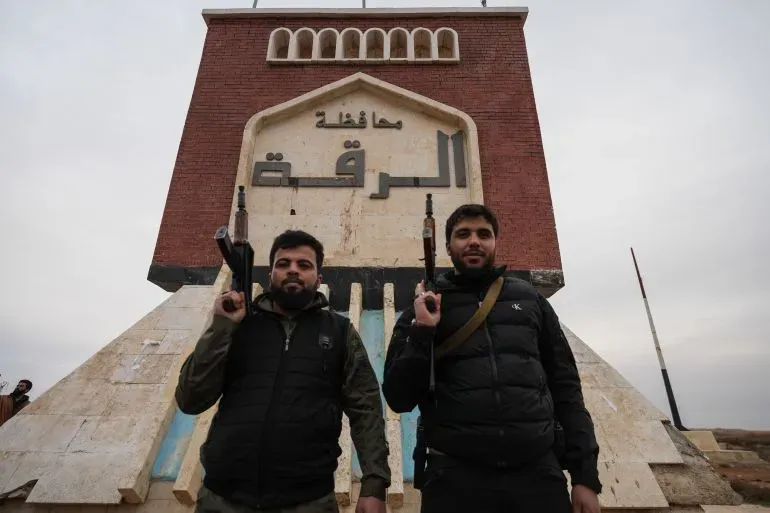 مقاتلان يحملان أسلحة يقفان أمام لافتة كبيرة مكتوبة عليها "محافظة الرقة"، في سياق السيطرة العسكرية للجيش السوري على المنطقة.