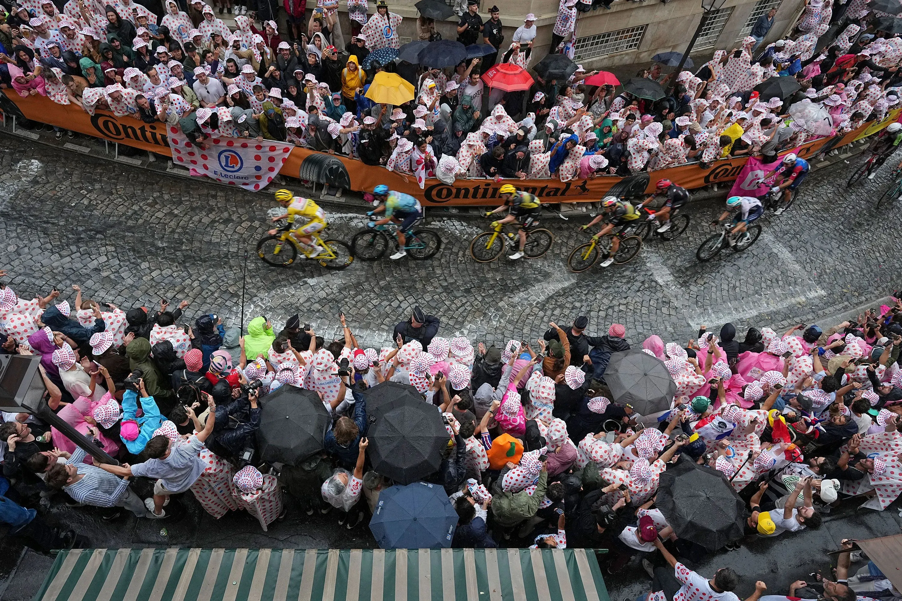 احتفال حشود كبيرة بسباق طواف فرنسا للدراجات، حيث يتسابق المتسابقون في الشوارع المبللة تحت الأمطار، مع ارتداء المشجعين قبعات مزينة.