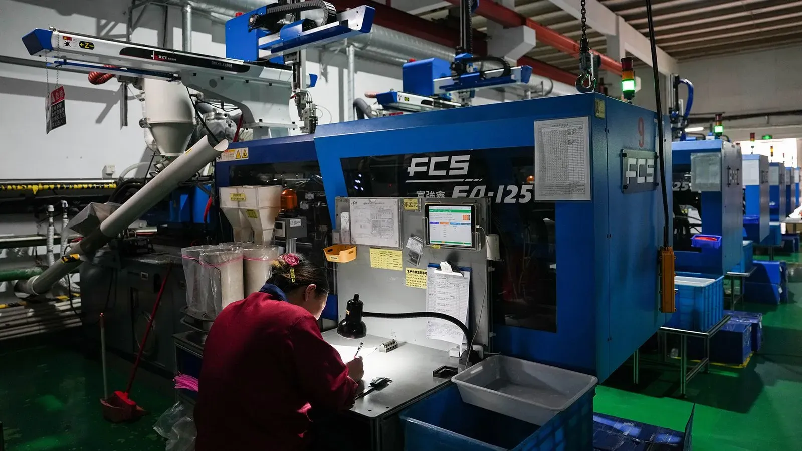 امرأة تعمل على آلة تصنيع في مصنع صيني، مع معدات متطورة وخطوط إنتاج، تعكس تأثير الحرب التجارية على الصناعة.