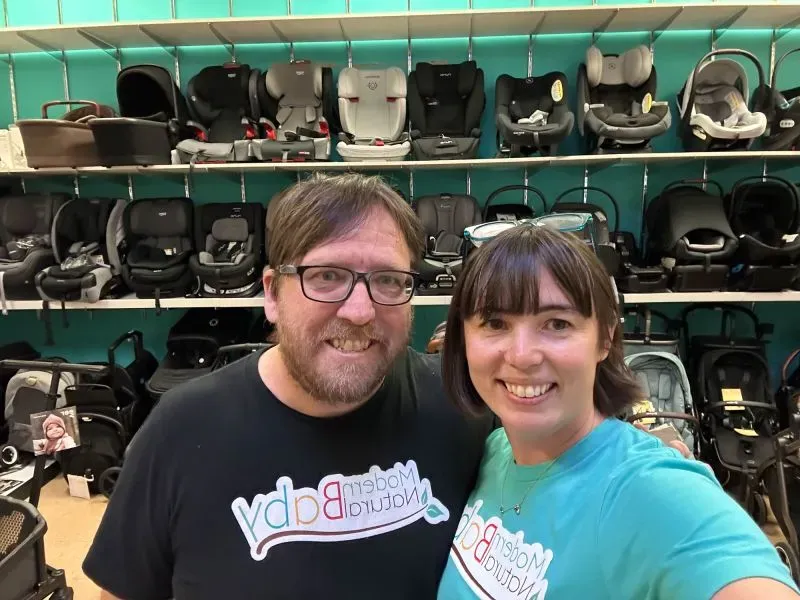 زوجان يبتسمان في متجر لمعدات الأطفال، مع خلفية تضم مجموعة متنوعة من عربات الأطفال ومقاعد السيارة، يعكسان قلق الآباء بشأن ارتفاع الأسعار.