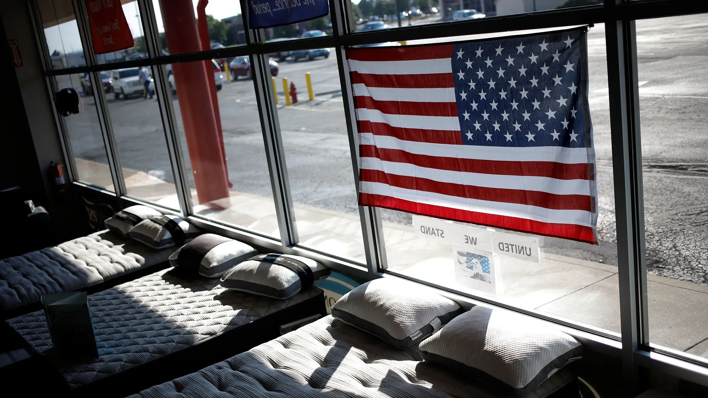 علم الولايات المتحدة يتدلى من نافذة متجر يبيع المراتب، مع وضع المراتب والوسائد في الداخل، مما يعكس تأثير التعريفات الجمركية على الأسعار.