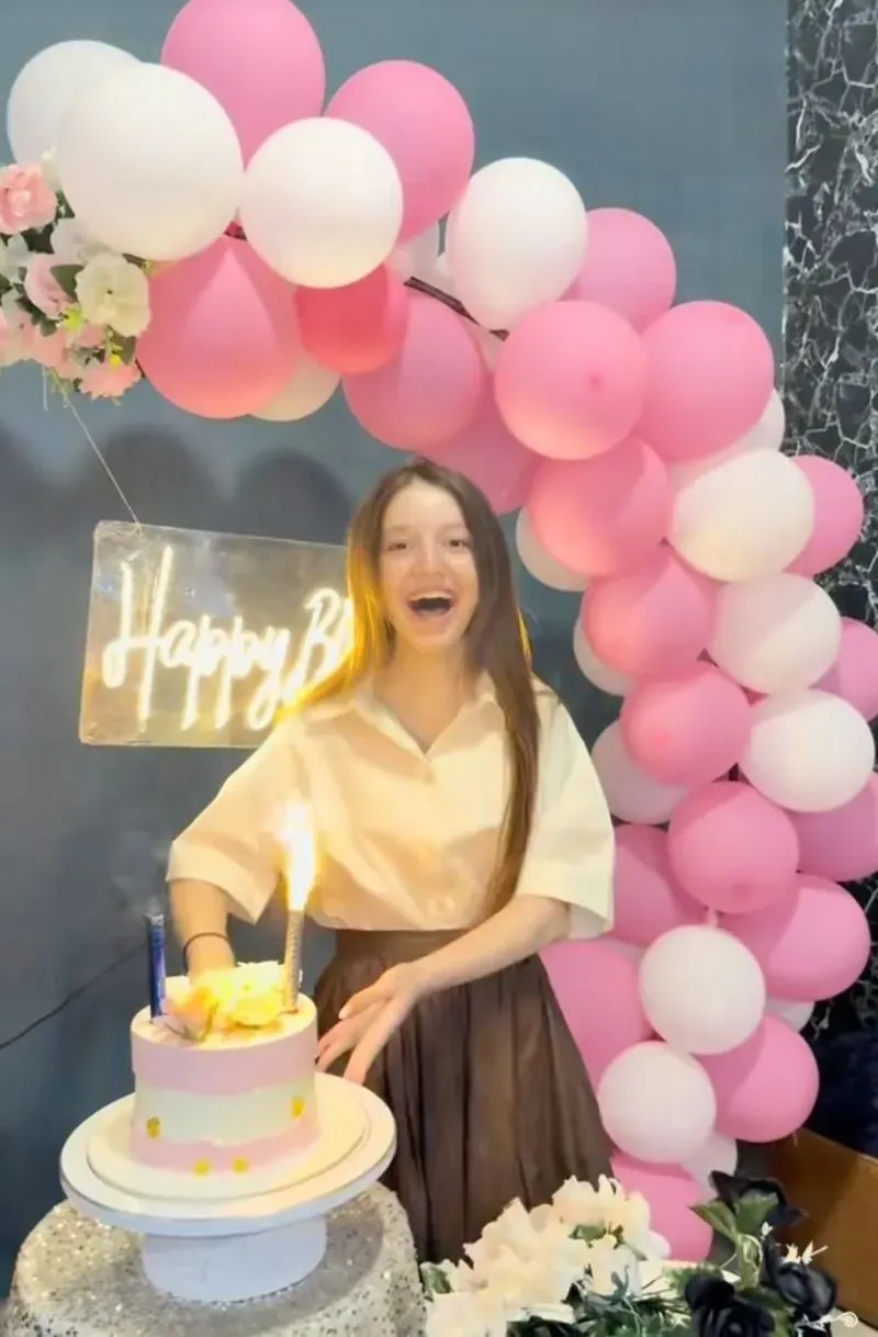 سناء يوسف تحتفل بعيد ميلادها السابع عشر، مبتسمة أثناء قطع كعكة وردية، مع قوس بالونات خلفها، في أجواء احتفالية.