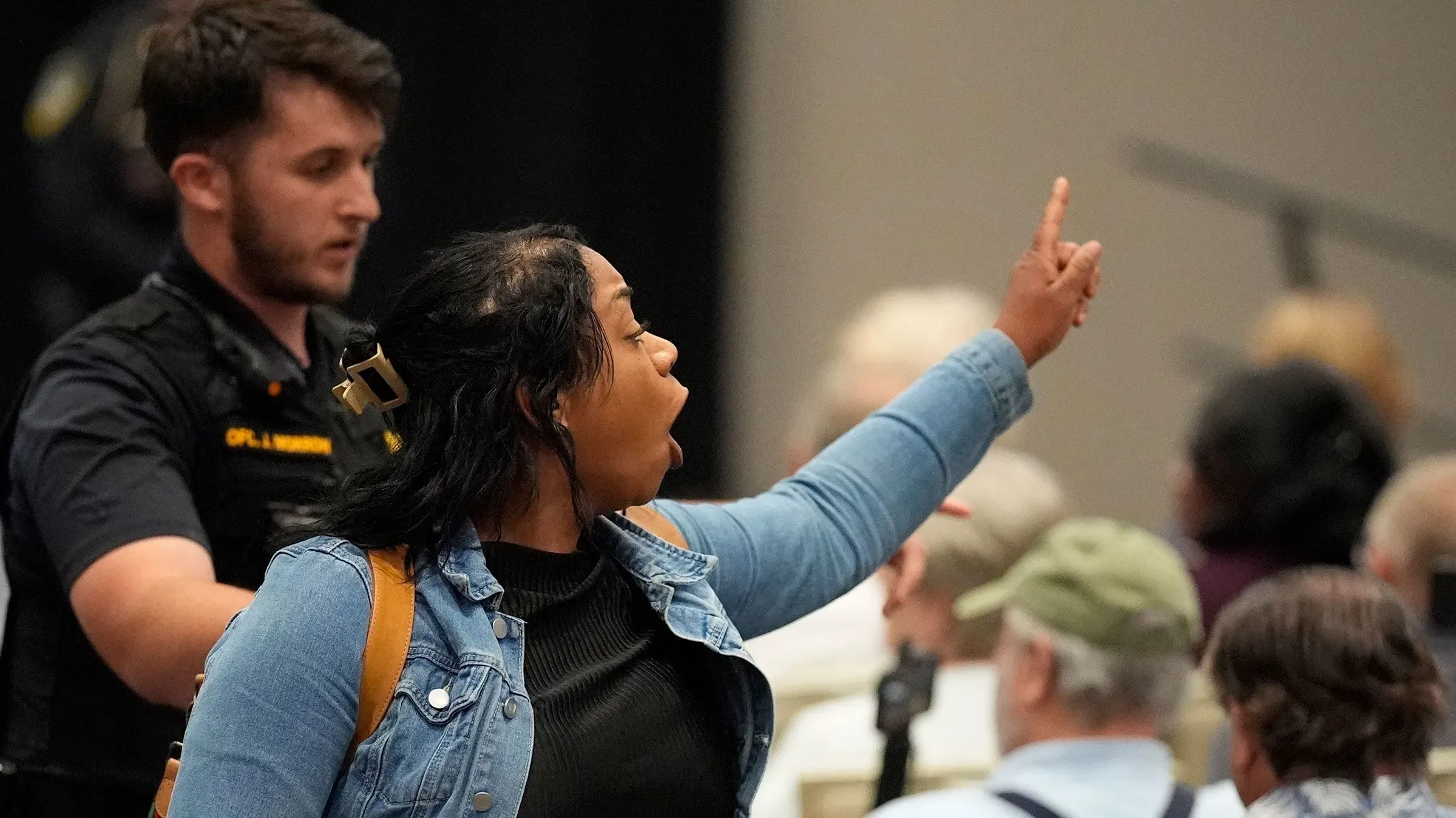 امرأة تعبر عن احتجاجها في اجتماع مجلس بلدية، بينما يقوم ضابط أمن بمرافقتها، مع وجود جمهور خلفها.