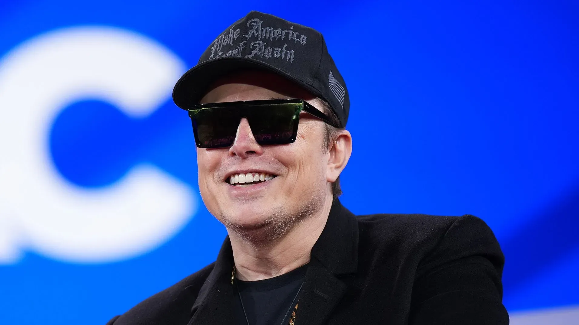 إيلون ماسك مبتسم أثناء حديثه، مرتديًا نظارات شمسية وقبعة، في سياق مناقشات حول تأثيره على مبيعات تسلا في أوروبا.