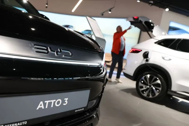 شعار شركة BYD على سيارة ATTO 3 سوداء، مع شخص في الخلفية يفتح صندوق سيارة بيضاء، مما يبرز المنافسة في سوق السيارات الكهربائية.