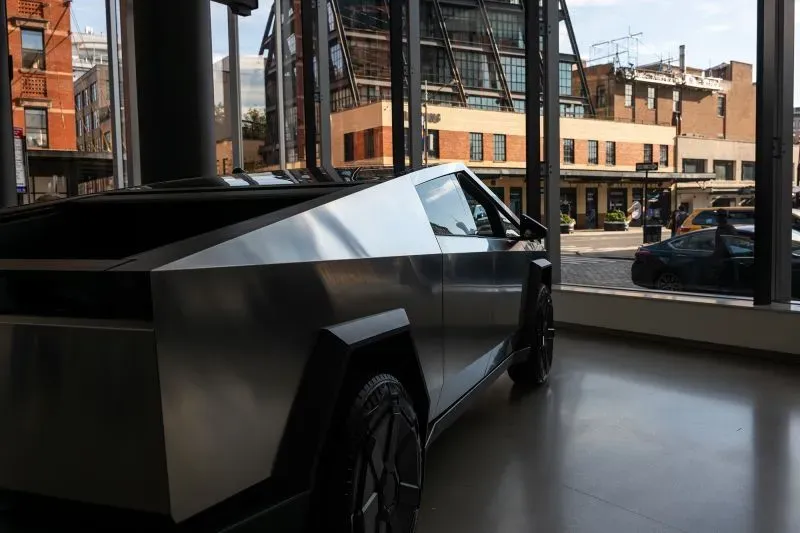 سيارة تسلا سايبر ترك ذات التصميم الهندسي الفريد متوقفة داخل صالة عرض، مع مشهد حضري من النوافذ الكبيرة في الخلفية.