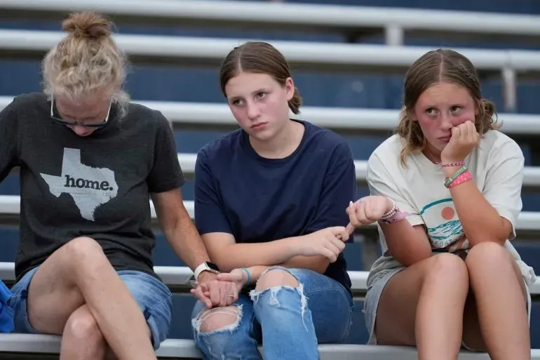 مجموعة من النساء الشابات يجلسن معًا في ملعب تيفي آنتلر، يعبرن عن الحزن خلال وقفة حداد على ضحايا الفيضانات في تكساس.