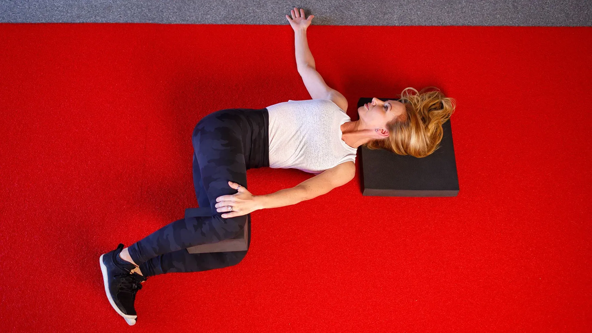 امرأة تؤدي تمرينًا على حصيرة حمراء، مستلقية على ظهرها مع ثني ركبتيها، ممسكة بوسادة بين ركبتيها، في سياق روتين لتحسين صحة العمود الفقري.