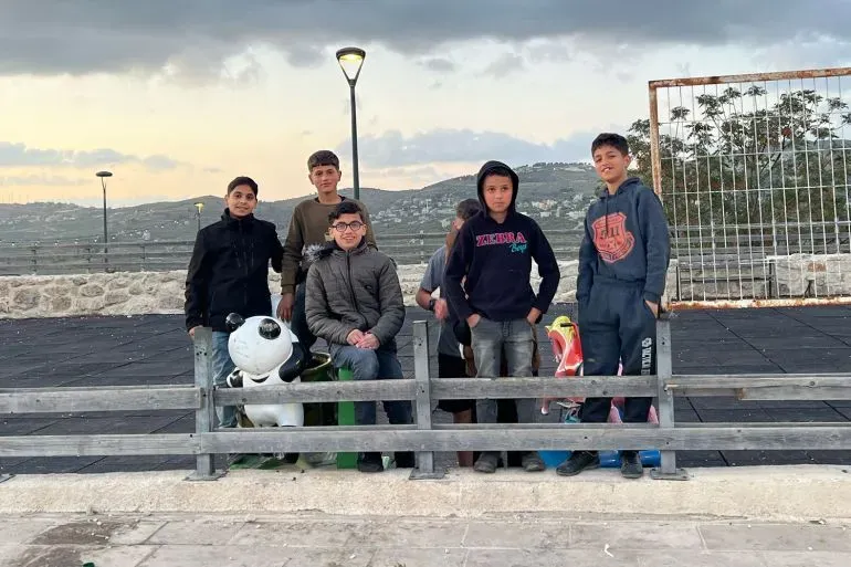 مجموعة من الأطفال والشباب في سبسطية، الضفة الغربية، يتجمعون في حديقة، في خلفية منظر طبيعي جبلي تحت سماء غائمة.