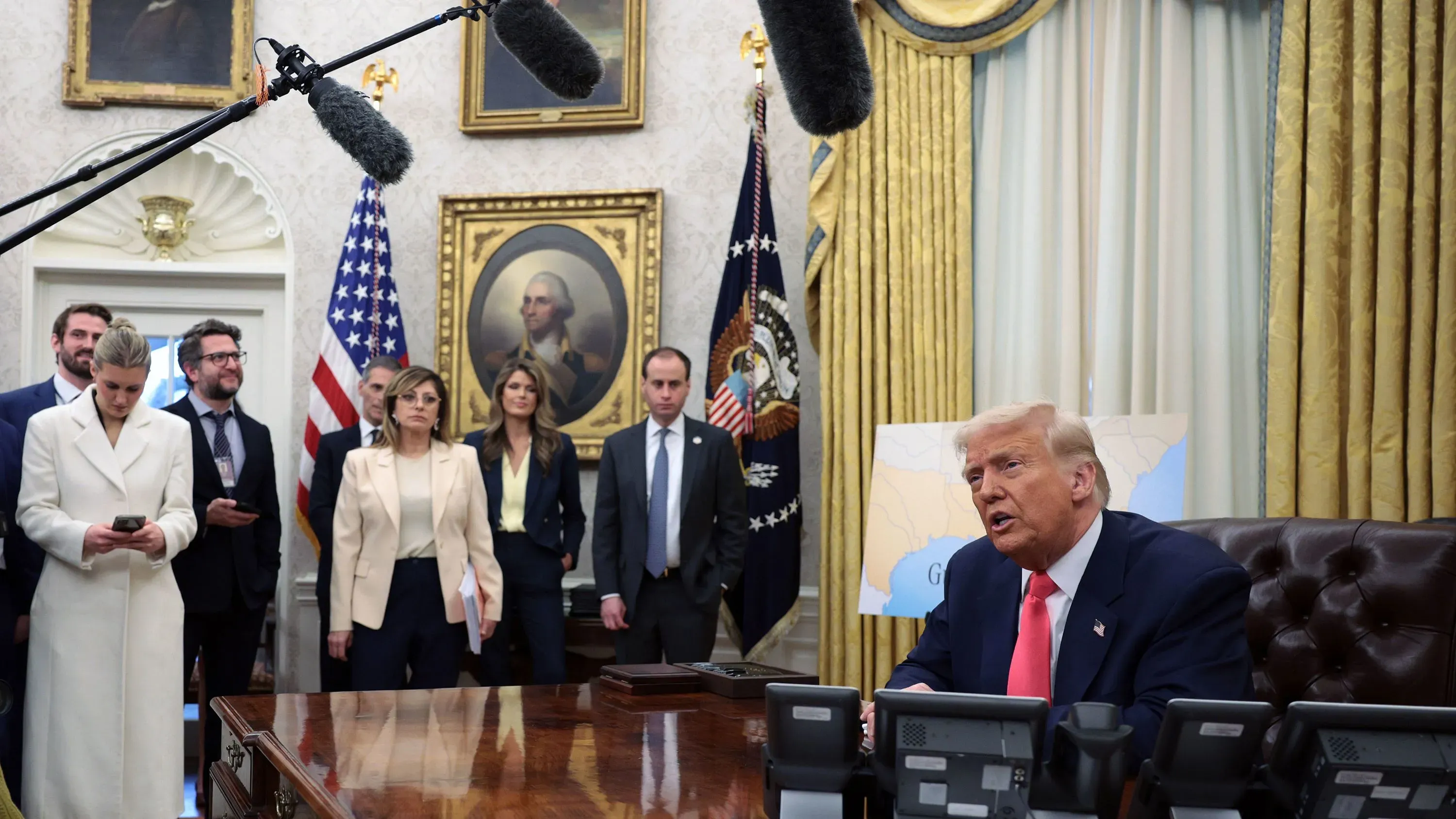 ترامب يتحدث في المكتب البيضاوي محاطًا بمساعديه، بينما تبرز أعلام الولايات المتحدة وصور تاريخية على الجدران، مما يعكس التوترات القانونية الحالية.