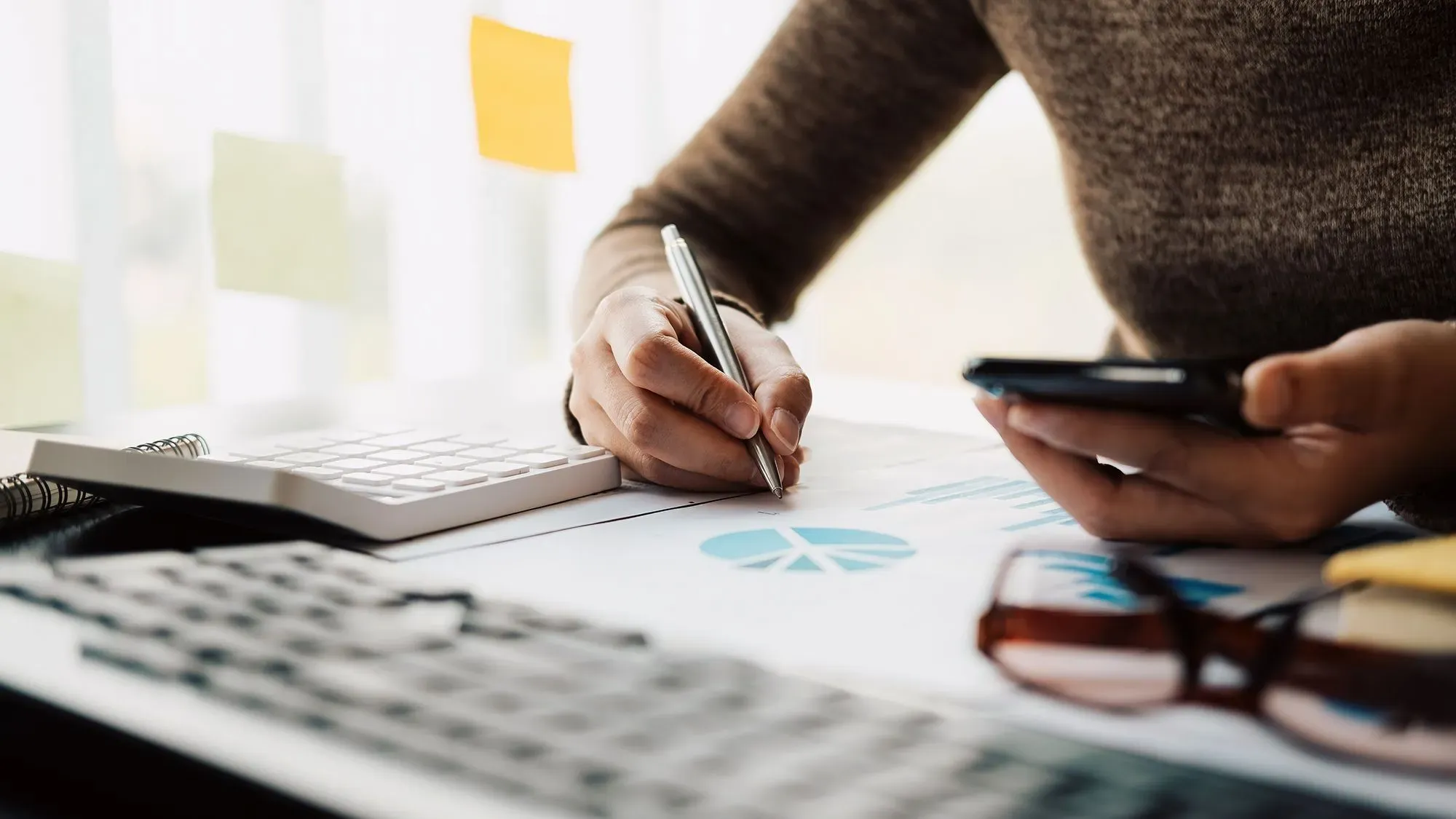 امرأة تستخدم قلمًا لتدوين الملاحظات على ورقة تحتوي على رسم بياني، بينما تمسك بهاتفها الذكي، مع وجود آلة حاسبة ونظارات على الطاولة.